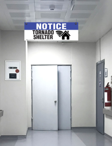 A double-sided emergency path sign printed with the words "Notice: Tornado Shelter" and a directional arrow, designed to hang from facility ceilings.